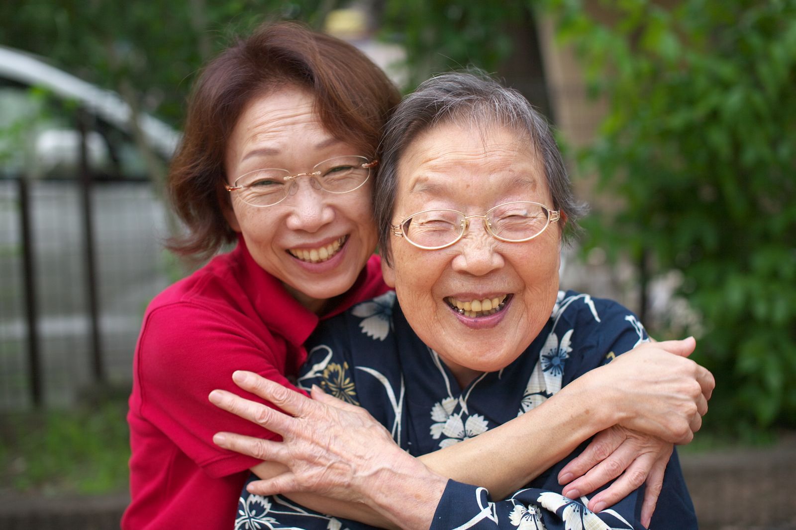 An older East Asian woman and her adult daughter hugging and smiling outdoors